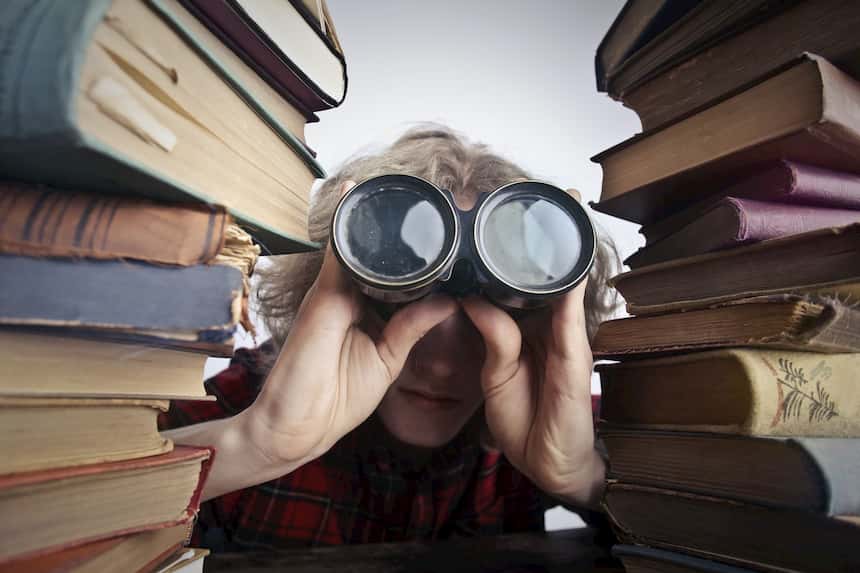 Person looking through binoculars surrounded by stacked books, symbolizing research and discovery