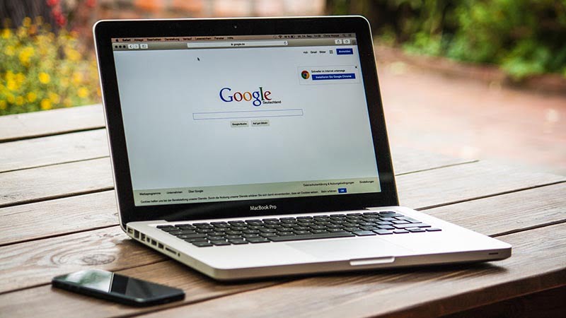 MacBook Pro displaying Google search homepage on outdoor wooden table with smartphone