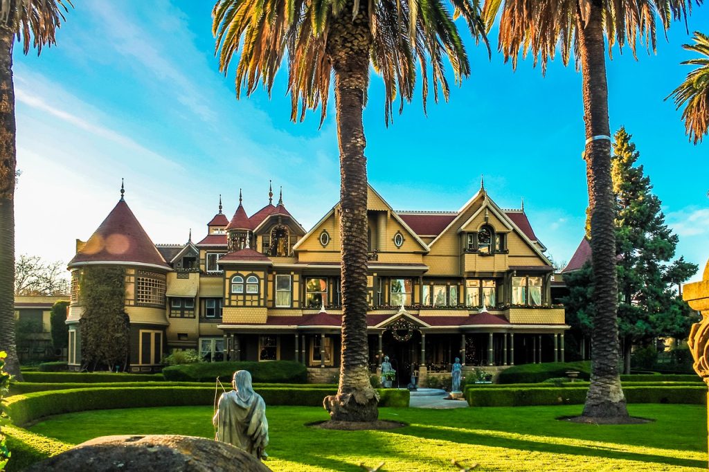 Victorian mansion with yellow exterior, red turrets, and manicured lawn framed by palm trees under blue sky