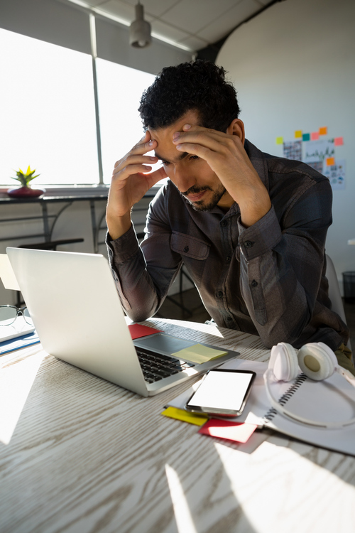Frustrated businessman at desk reviewing laptop screen with head in hands