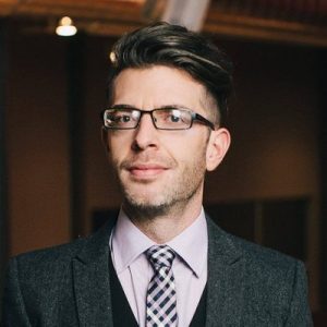 Professional headshot of man wearing glasses, gray blazer, and checkered tie