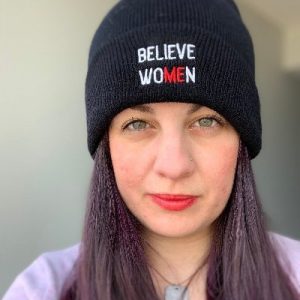 Person wearing dark beanie with "BELIEVE WOMEN" text and red accents