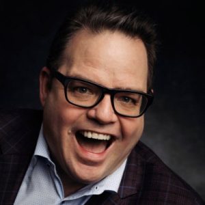 Man with dark hair and glasses wearing plaid blazer and light blue shirt, smiling at camera