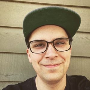 Joel Klettke smiling at camera wearing glasses and green cap