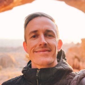 Man in black jacket smiling outdoors with desert landscape background