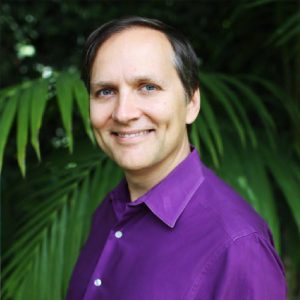 Digital marketing professional in purple shirt smiling outdoors with green foliage