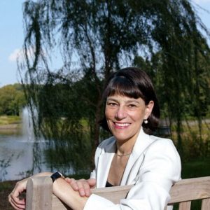Veronica Stoddart smiling outdoors by a wooden railing with weeping willow and pond background