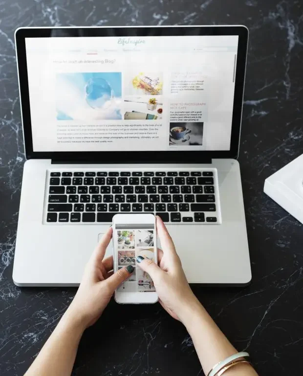 Hands holding smartphone displaying social media feed above laptop showing website design