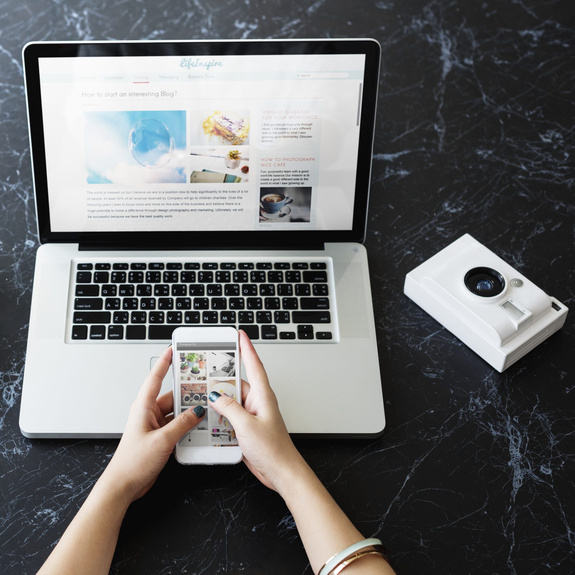 Hands holding smartphone displaying social media feed, with laptop showing lifestyle blog website and white camera on marble