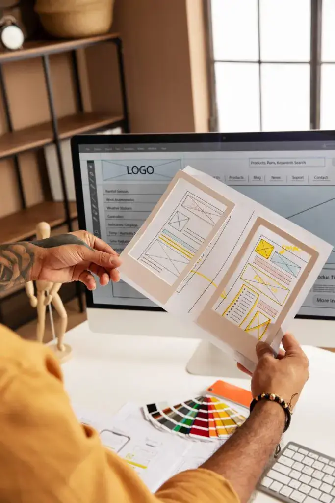 Designer reviewing website wireframe sketches with color palette and monitor displaying logo design