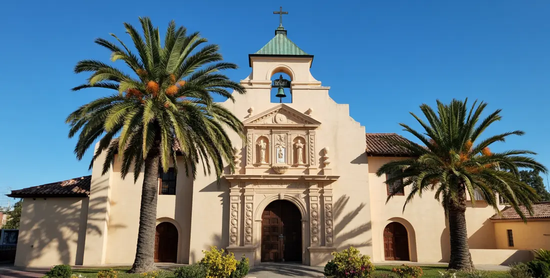 Historic Spanish mission church with cream stucco facade, bell tower, cross, palm trees under clear blue sky