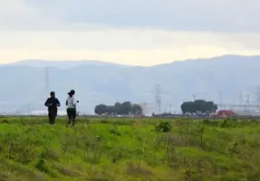 Two runners jogging through Bedwell Bayfront Park with San Francisco Bay Area hills in the background