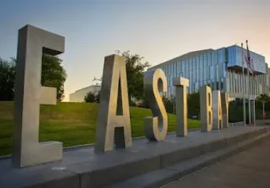 Large "HAYWARD" letters monument with modern building on grassy hilltop at sunset