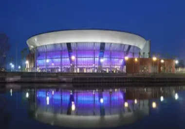 Modern arena with illuminated blue and purple glass facade reflected in water at dusk
