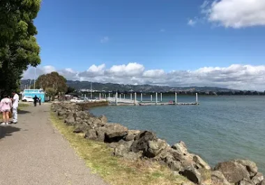 Waterfront path with rocky shoreline and sailboats on bay under blue sky